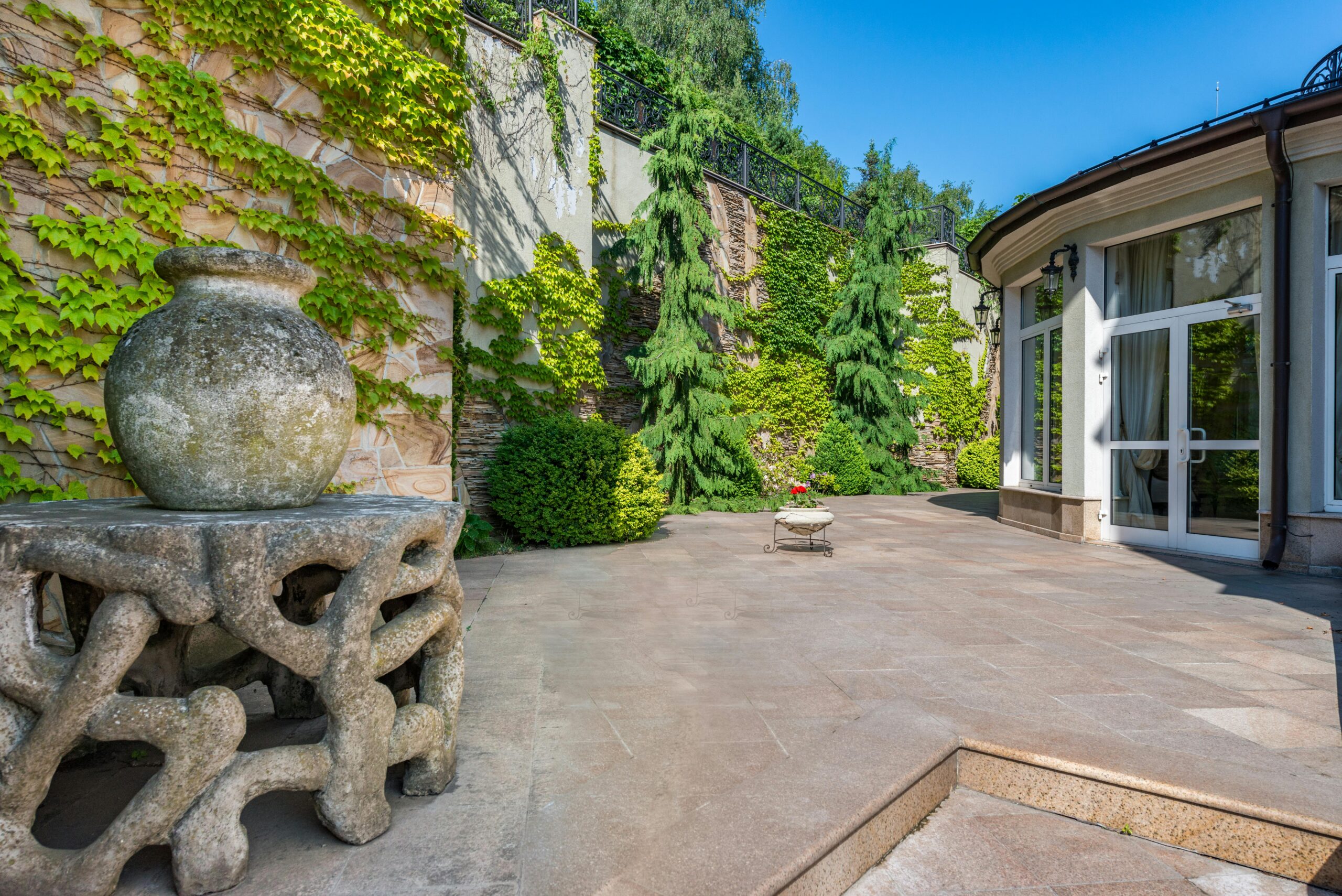 Exterior of house with large windows placed in spacious yard with aged stone sculptures against high wall covered with green plants in daytime
